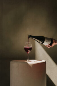 In ein Weinglas wird elegant Rotwein eingeschenkt. Das Etikett auf der Weinflasche zeigt das Winzer Logo.